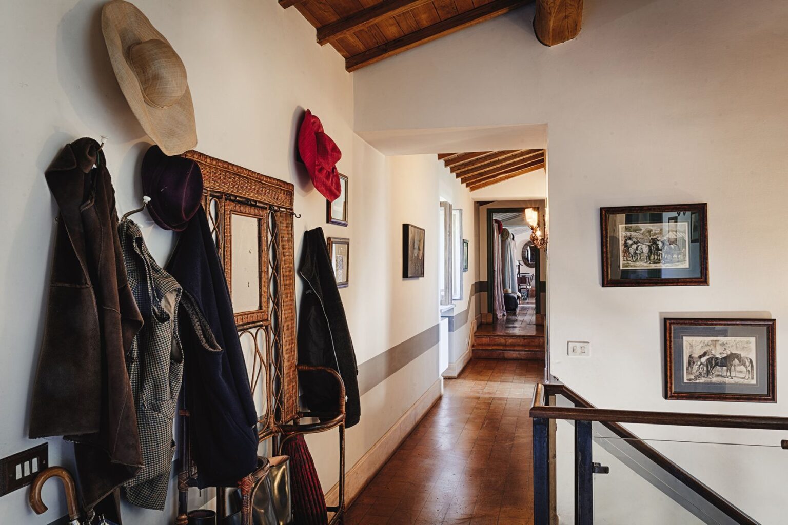 Interior hallway featuring a wooden ceiling, displaying various hats and jackets on a wall-mounted rack, along with framed artwork. The corridor leads to another room with wooden stairs and additional decor. | Only on SopranoVillas
