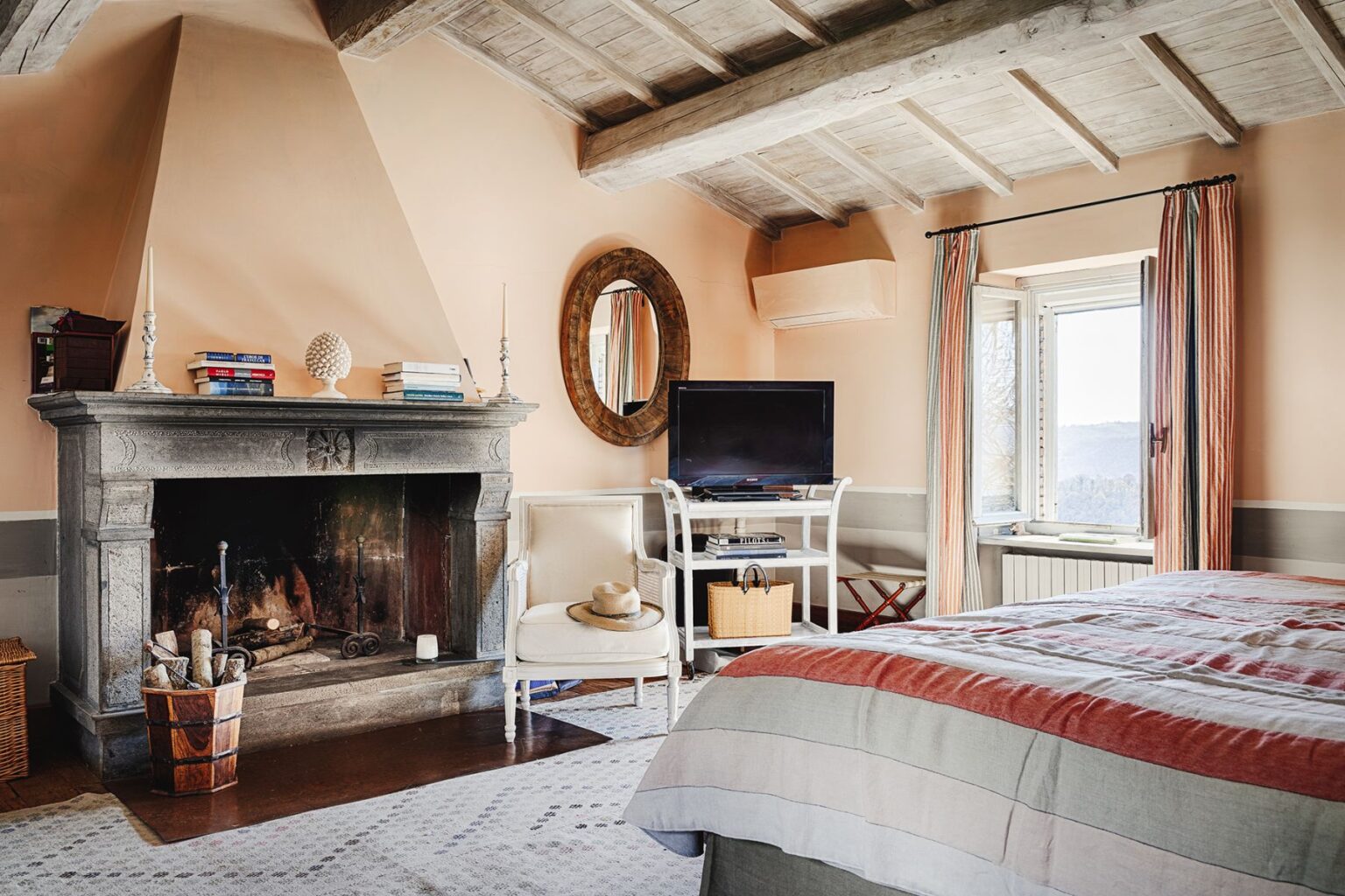 Cozy bedroom featuring a rustic stone fireplace, a large window with striped curtains, and a comfortable bed with striped bedding. The room is decorated in soft peach tones, with a round mirror, books on the mantel, and a TV on a stylish stand, creating a warm and inviting atmosphere. | Only on SopranoVillas
