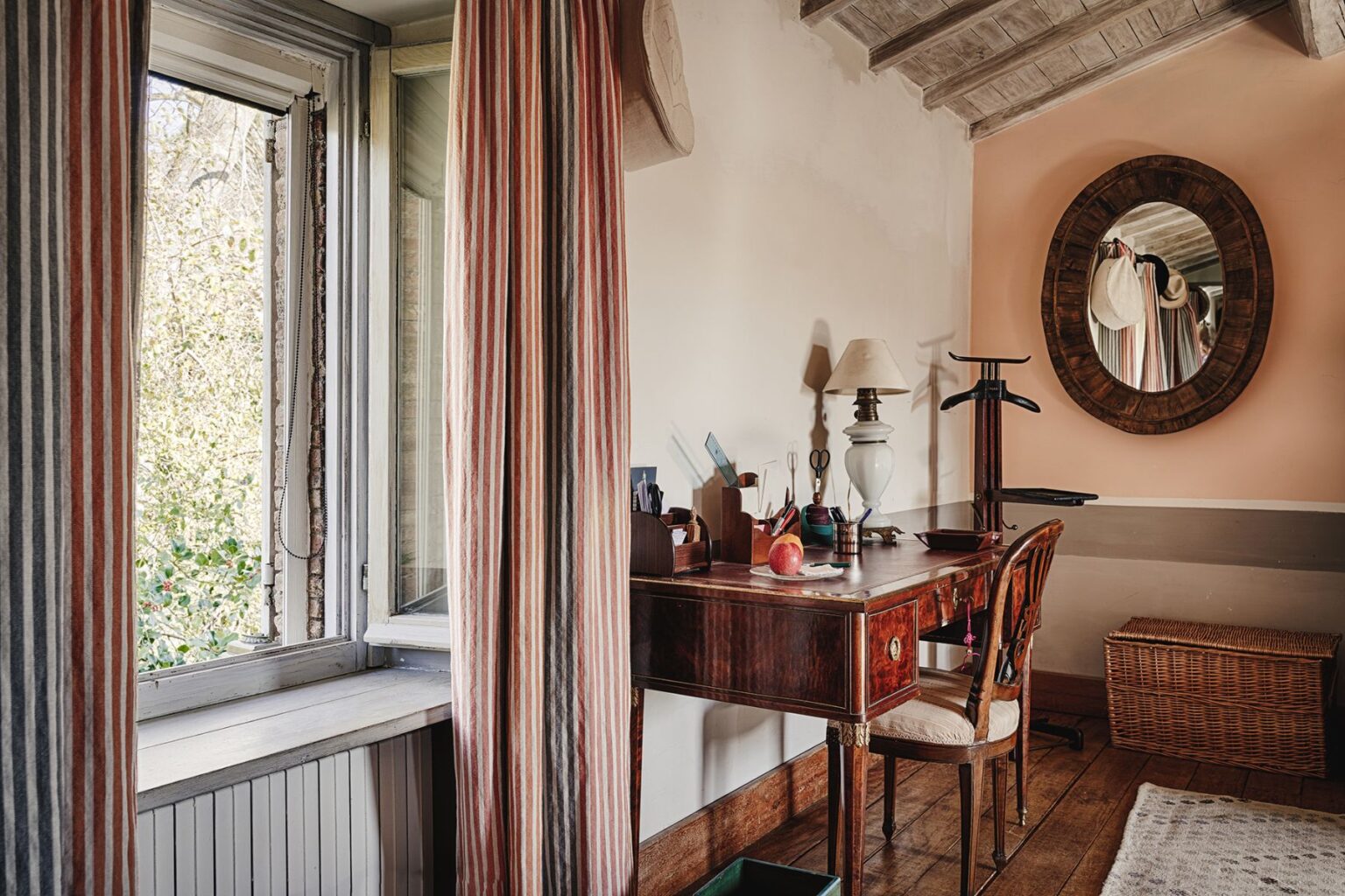 Cozy interior of a home office featuring a wooden desk with stationery, an apple, and a lamp, complemented by striped curtains and a mirror. Natural light streams in through the window, enhancing the warm ambiance. | Only on SopranoVillas
