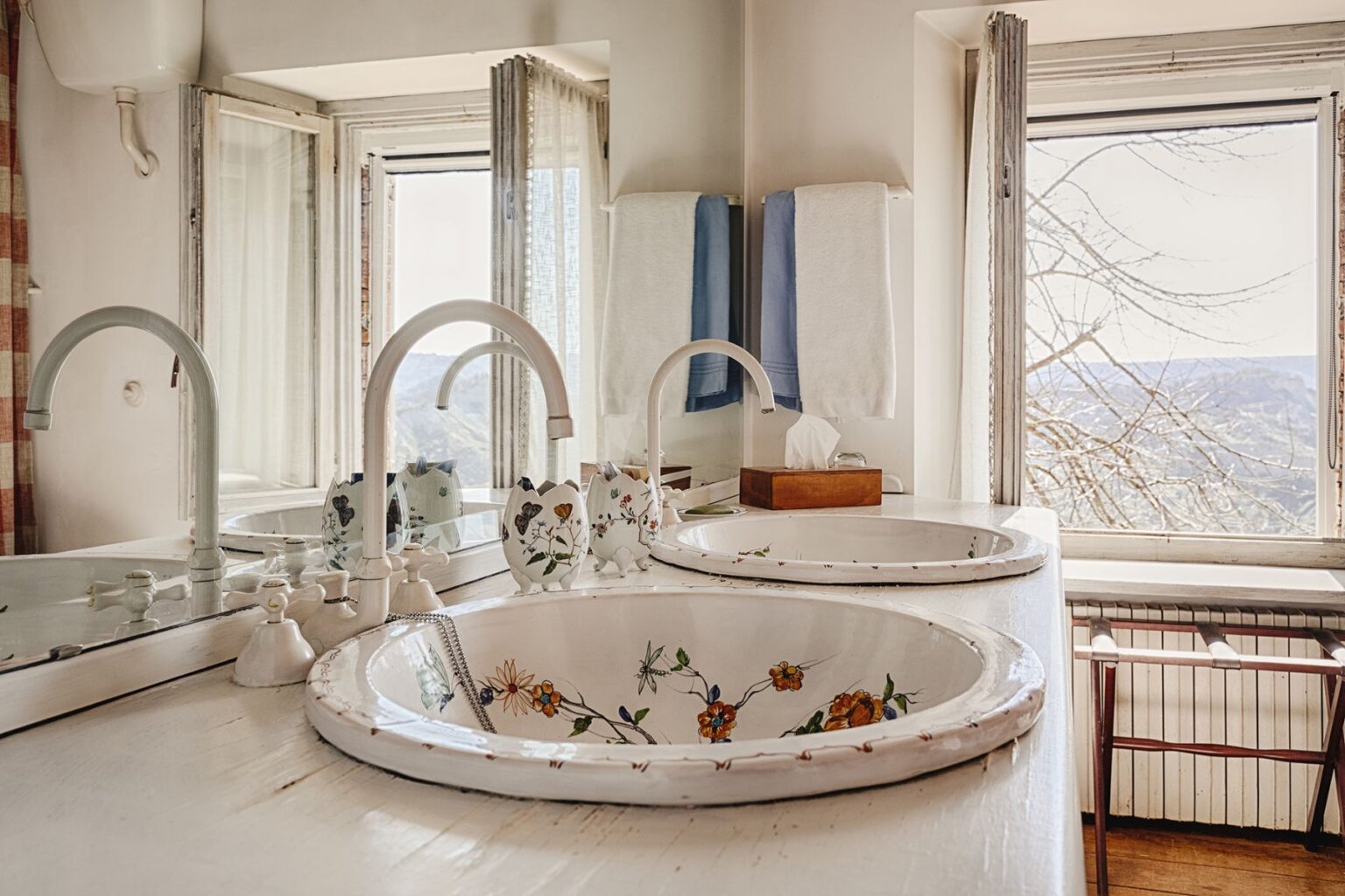 Bright and airy bathroom featuring two vintage sinks with floral designs, modern faucets, and a scenic view through large windows. Soft towels hang nearby, enhancing the tranquil atmosphere. | Only on SopranoVillas