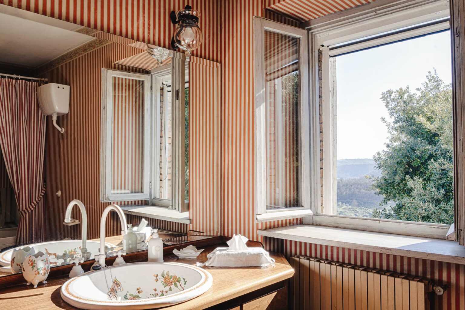 Bright and stylish vintage bathroom featuring a floral sink, wooden countertop, and striped wallpaper. Large windows provide a scenic view of the landscape, enhancing the room's natural light. | Only on SopranoVillas