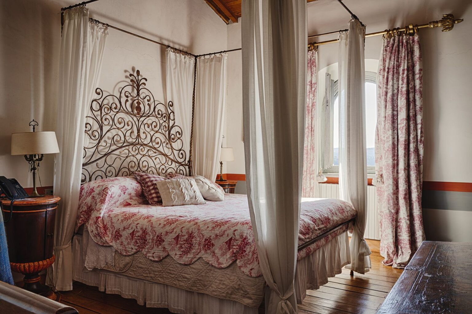 Cozy bedroom featuring an ornate wrought iron bed frame with floral bedding, sheer white drapes, and elegant curtains. The room includes wooden flooring, a bedside lamp, and a decorative side table, creating a warm and inviting atmosphere. Natural light streams in through a window, enhancing the serene ambiance. | Only on SopranoVillas