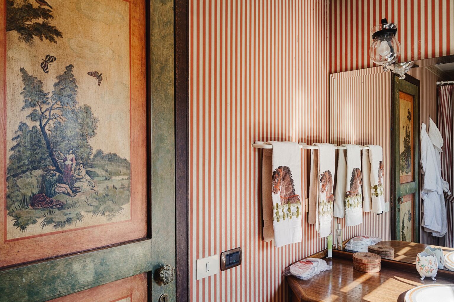 Vintage bathroom interior featuring striped wallpaper and a decorative door with a pastoral scene. The space includes neatly arranged towels with horse motifs, wooden vanity, and natural light illuminating the details. Ideal for design inspiration and home decor enthusiasts. | Only on SopranoVillas