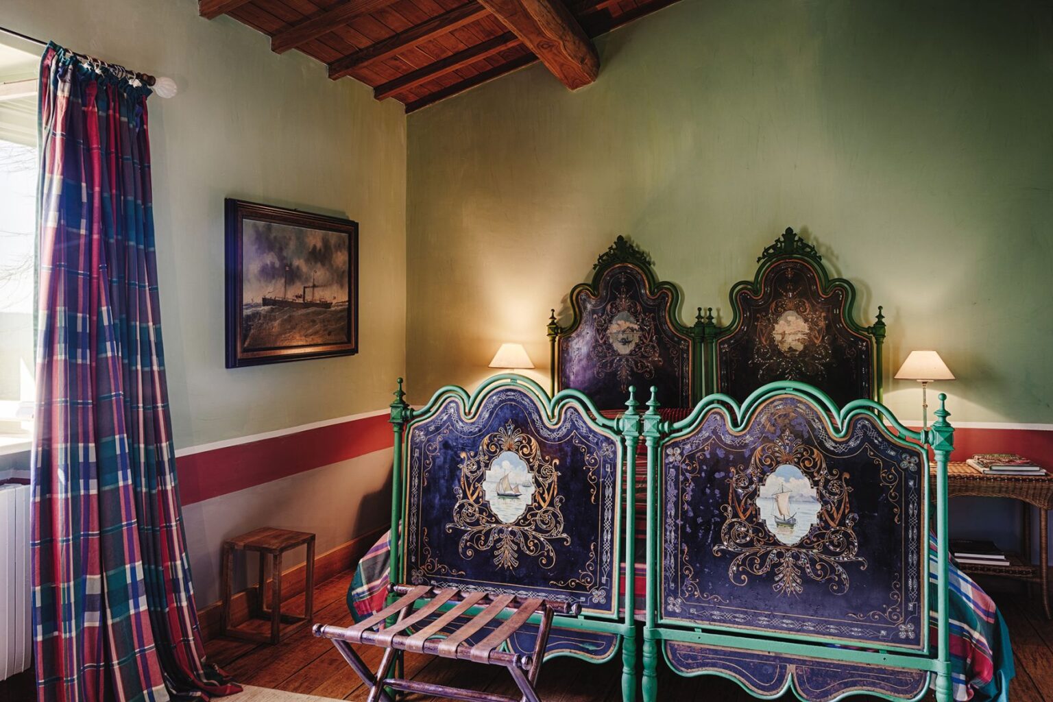 Cozy bedroom interior featuring ornate green and blue vintage twin beds with intricate designs, a plaid curtain, a wooden chair, and a framed painting on the wall, illuminated by a soft bedside lamp. | Only on SopranoVillas