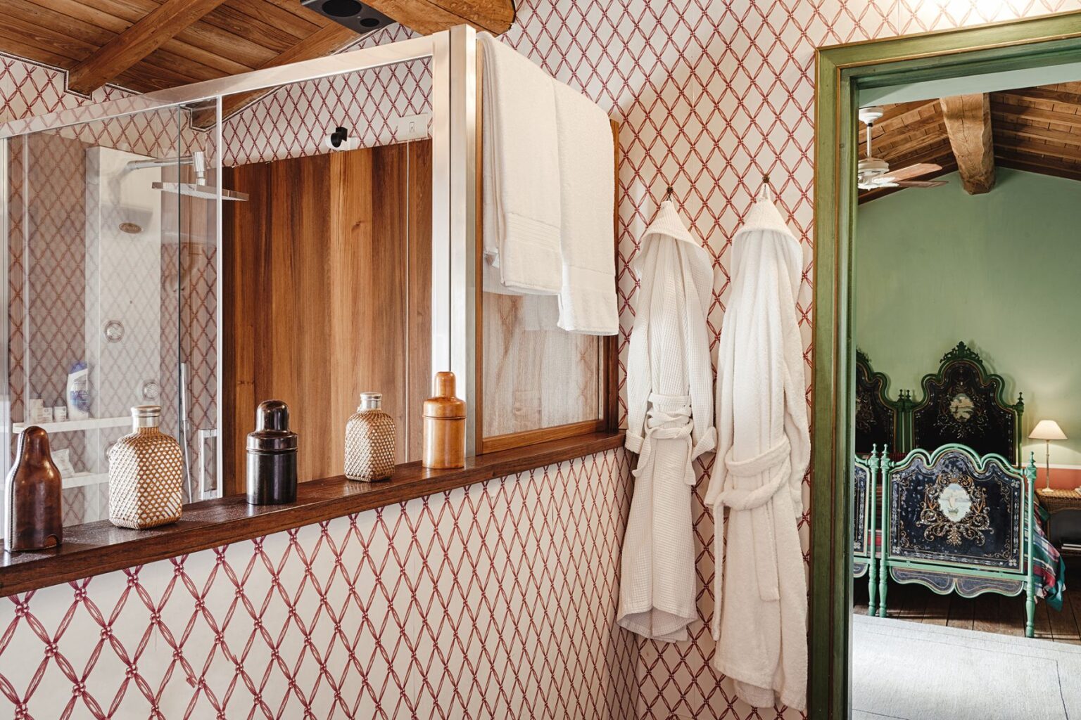 Modern bathroom interior featuring a glass shower, wooden accents, and decorative bottles on a shelf. White bathrobes hang on the wall, with stylish patterned wallpaper and a glimpse of a bedroom with ornate furniture visible through the doorway. | Only on SopranoVillas