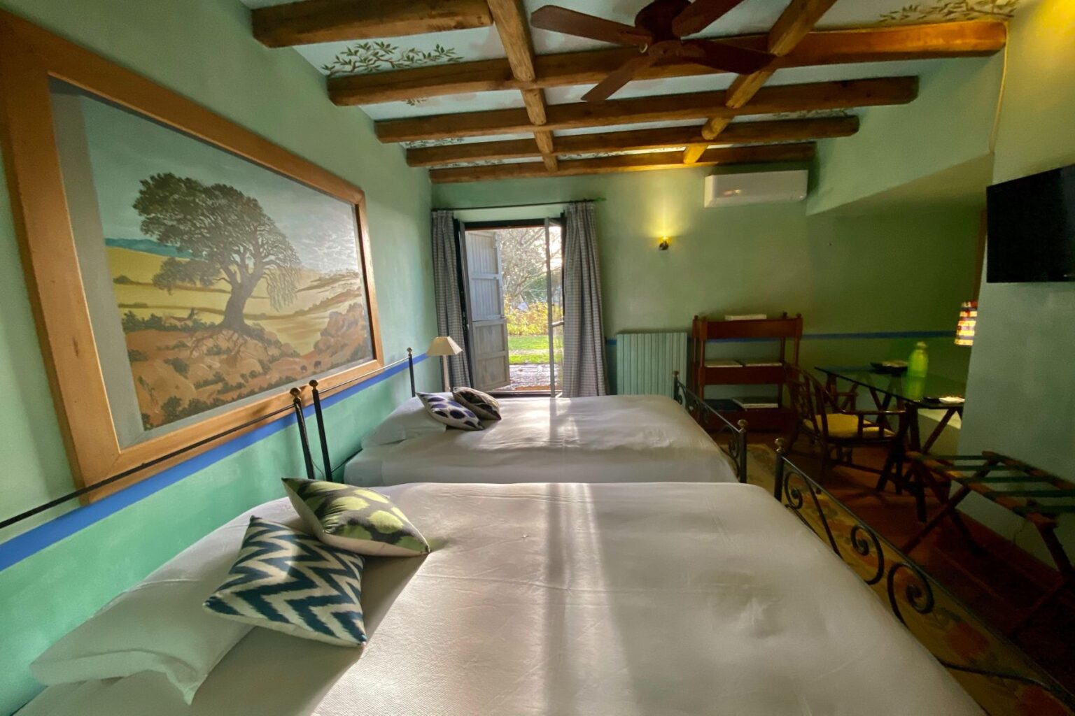 Cozy guest room featuring two beds with decorative pillows, a large artwork of a tree on the wall, wooden beams on the ceiling, and a view of a garden through an open door. The room is painted in soft green tones and includes a small table and a TV. Ideal for a relaxing stay. | Only on SopranoVillas