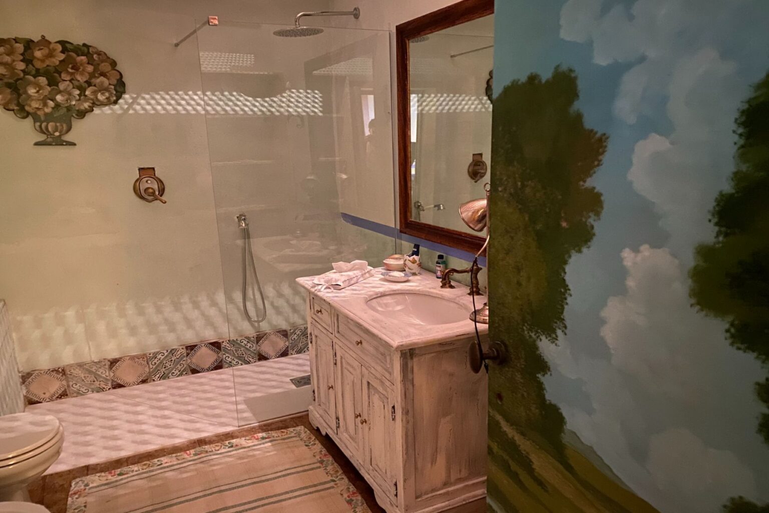 Bright and spacious bathroom featuring a glass shower, vintage-style sink with brass fixtures, and decorative wall art. The room includes tiled flooring and a cozy rug, creating a welcoming atmosphere. | Only on SopranoVillas