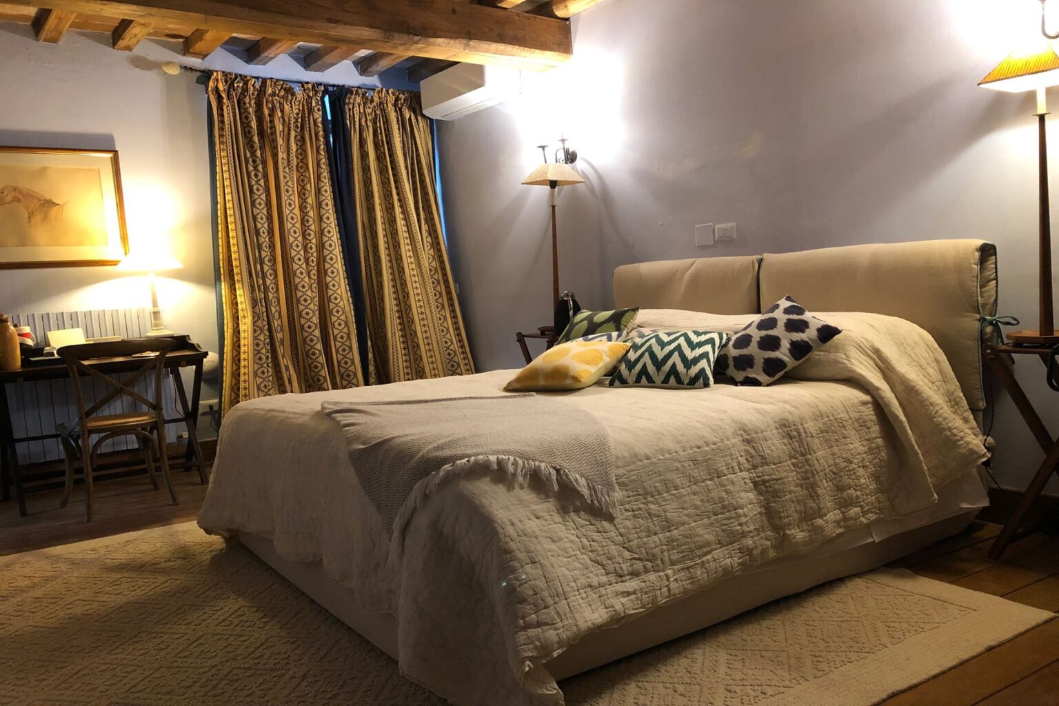 Cozy bedroom featuring a comfortable bed with decorative pillows, warm lighting from a bedside lamp, and elegant patterned curtains. A wooden desk is positioned nearby, enhancing the inviting atmosphere of the space. | Only on SopranoVillas