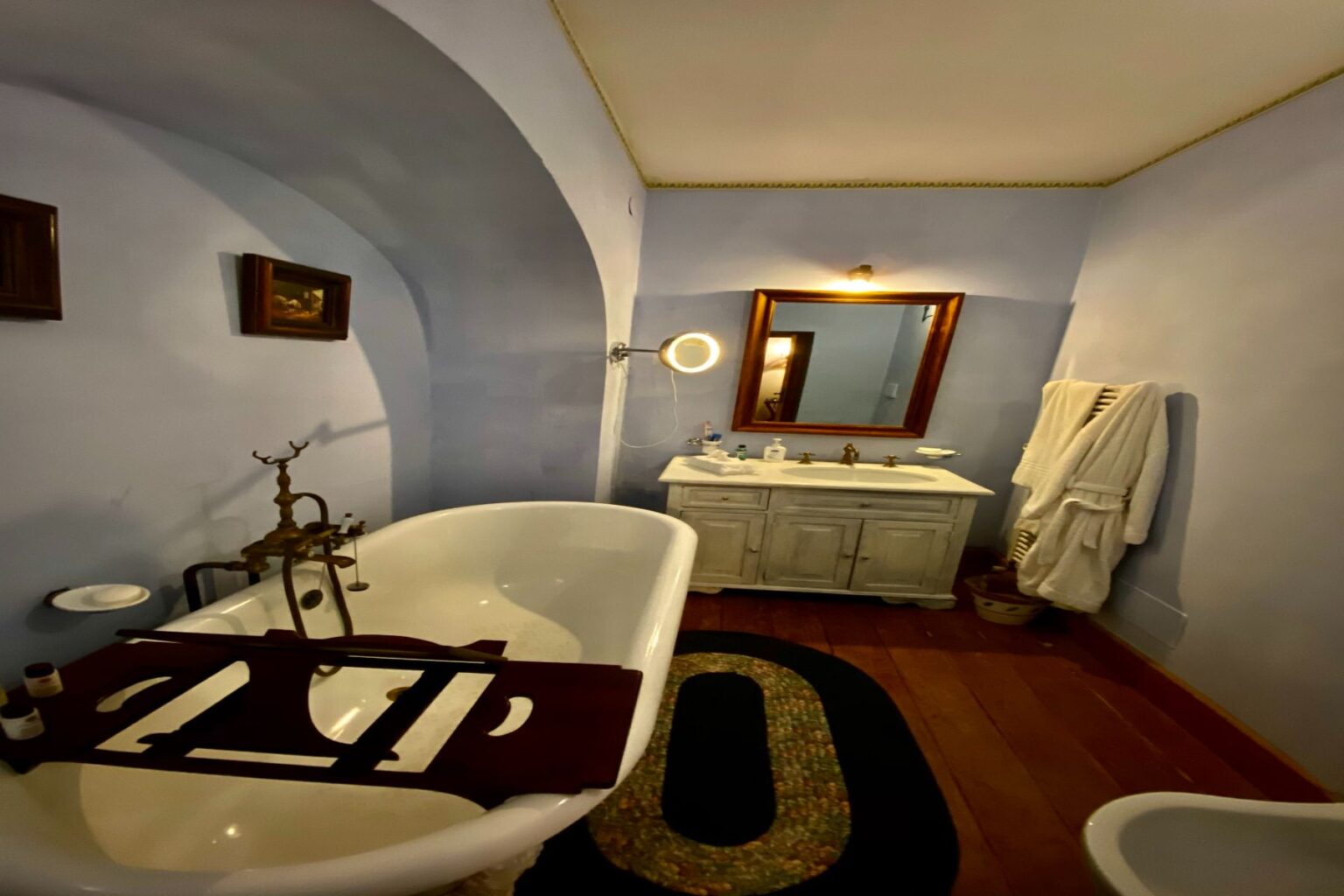 Bright and stylish bathroom featuring a vintage freestanding bathtub, a modern vanity with a mirror, and decorative wall art. The space is accented by soft blue walls, wooden flooring, and plush bathrobes, creating a cozy and inviting atmosphere. | Only on SopranoVillas