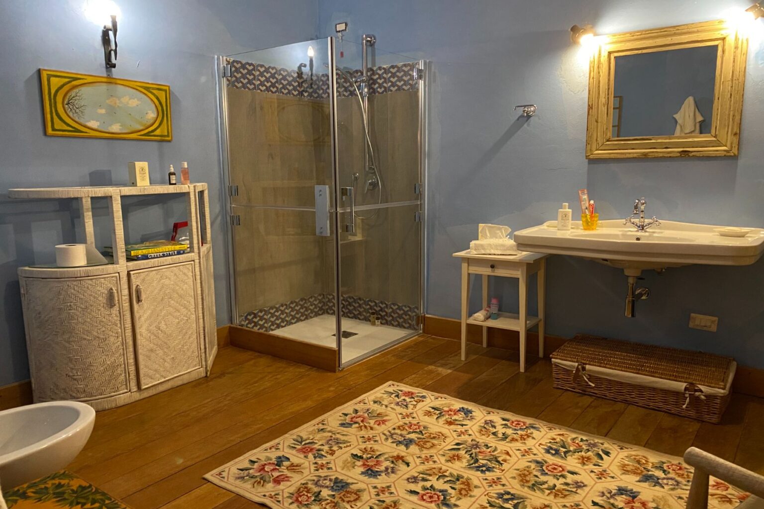 Bright and airy bathroom featuring a glass shower enclosure, white sink, and stylish decor. The space includes a decorative mirror, a patterned area rug, and a storage cabinet with toiletries. Warm wooden flooring complements the blue walls, creating a cozy atmosphere. | Only on SopranoVillas