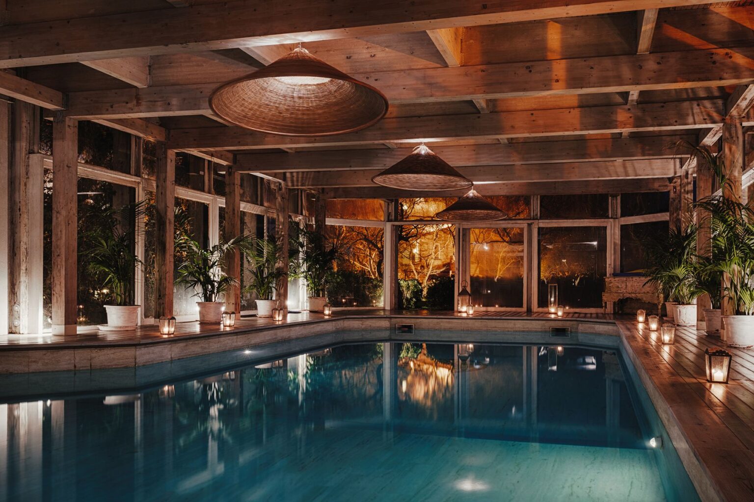 Indoor swimming pool with wooden beams and large windows, surrounded by potted plants and lanterns, illuminated softly for a relaxing ambiance at night. | Only on SopranoVillas