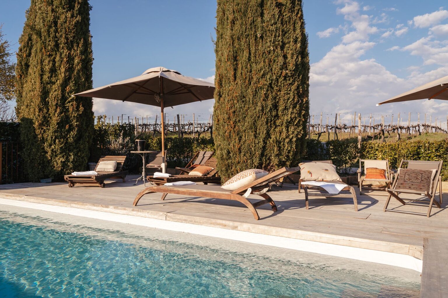 Lounge chairs and umbrellas by a tranquil swimming pool surrounded by lush greenery and cypress trees, with a scenic vineyard in the background under a clear blue sky. | Only on SopranoVillas
