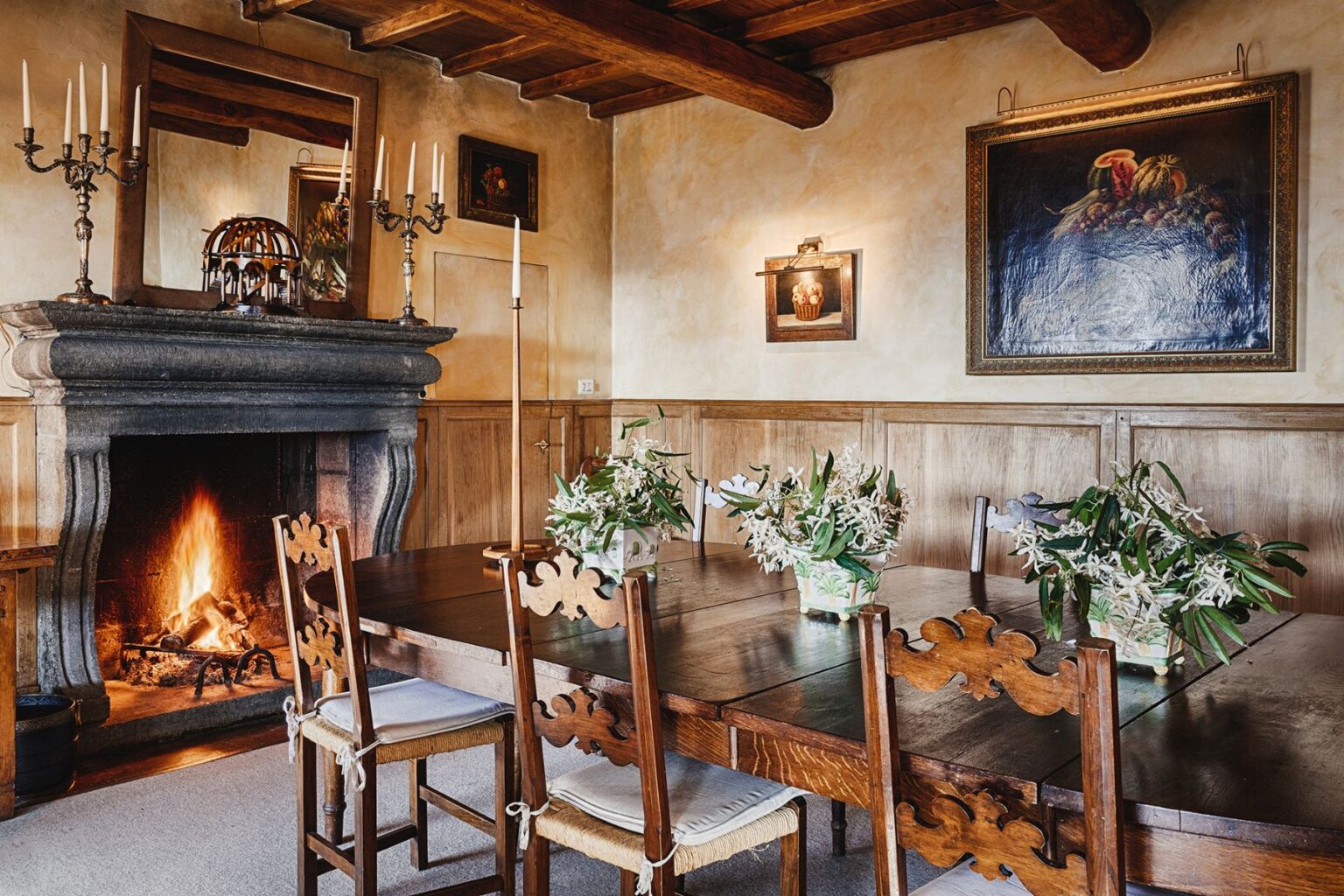 Cozy dining room featuring a rustic wooden table surrounded by ornate chairs, elegant floral arrangements, and a stone fireplace with a warm fire. The walls are adorned with artwork, and soft candlelight enhances the inviting atmosphere. | Only on SopranoVillas