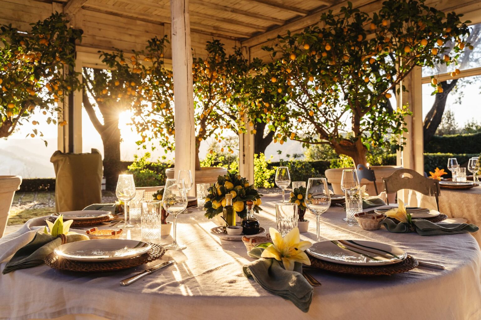 Elegant outdoor dining setup at sunset, featuring a beautifully arranged table with fine dinnerware, glassware, and floral centerpieces surrounded by lush citrus trees. Ideal for special events or intimate gatherings in a picturesque setting. | Only on SopranoVillas