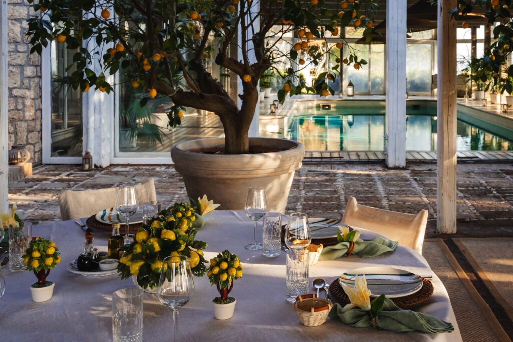 Elegant outdoor dining setup featuring a beautifully arranged table with lemon centerpieces, glassware, and plates, set against a backdrop of a serene swimming pool and a lush lemon tree. The setting exudes a relaxed, sophisticated atmosphere, perfect for gatherings. | Only on SopranoVillas
