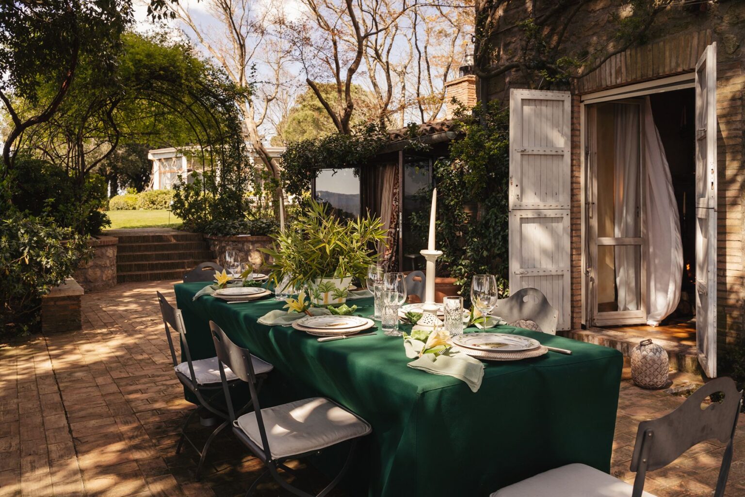 Outdoor dining setup featuring a long green table adorned with elegant tableware, a white candle, and decorative plants. The scene is surrounded by lush greenery and a charming stone pathway leading to a serene garden. Natural light filters through trees, enhancing the inviting atmosphere of the outdoor dining space. | Only on SopranoVillas