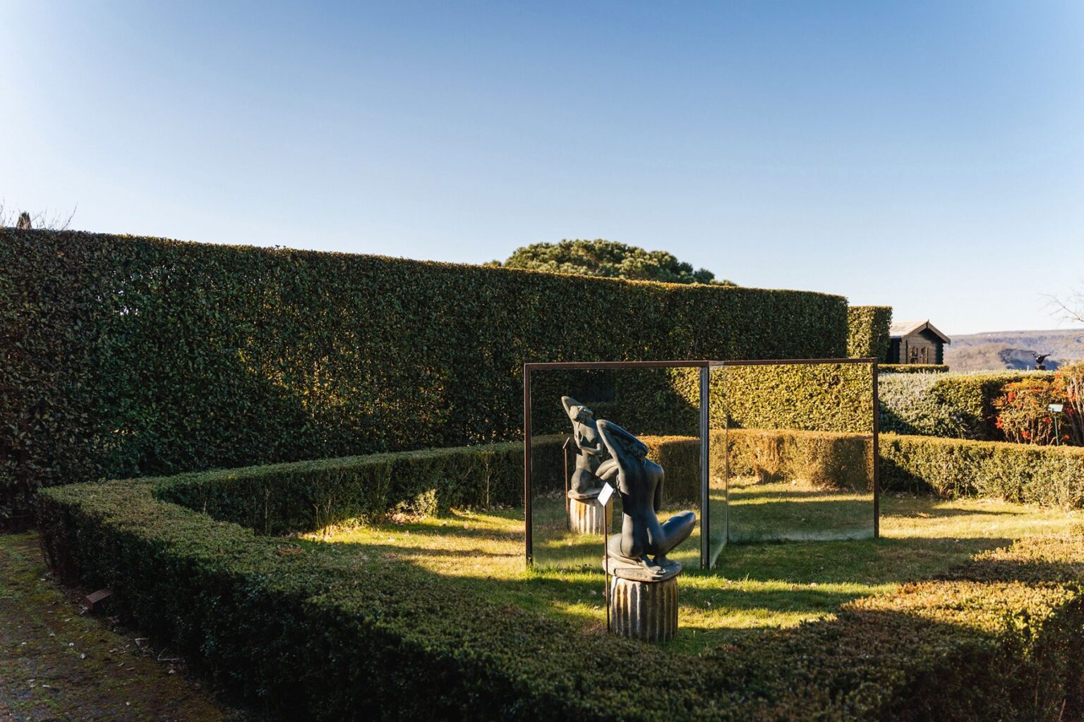 Sculpture surrounded by neatly trimmed hedges in a garden, featuring mirrors that reflect the artwork against a clear blue sky. | Only on SopranoVillas