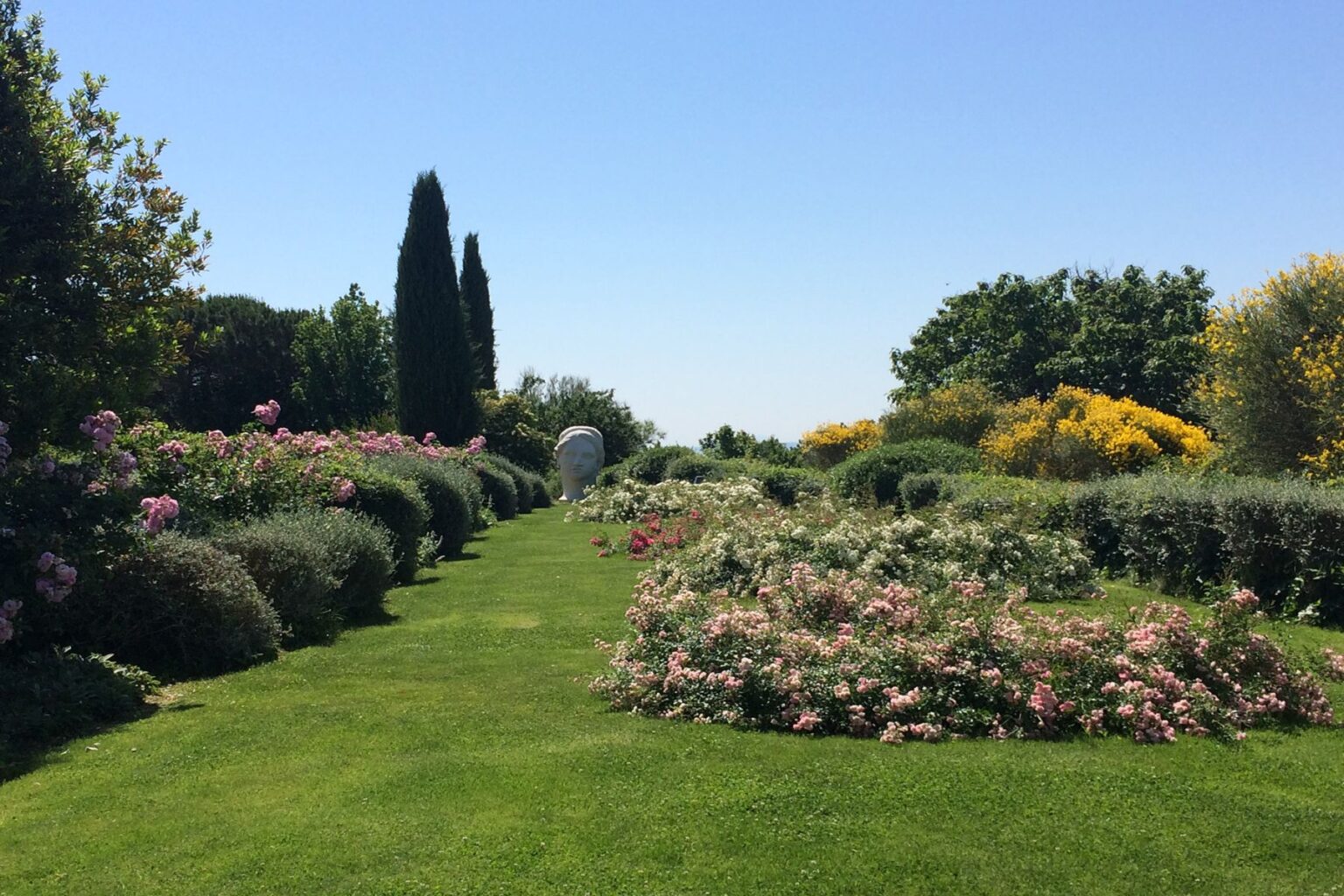 Lush garden pathway lined with colorful flowering shrubs, including pink roses and white blooms, framed by tall trees under a clear blue sky. A white sculpture is visible in the distance, enhancing the serene landscape. | Only on SopranoVillas