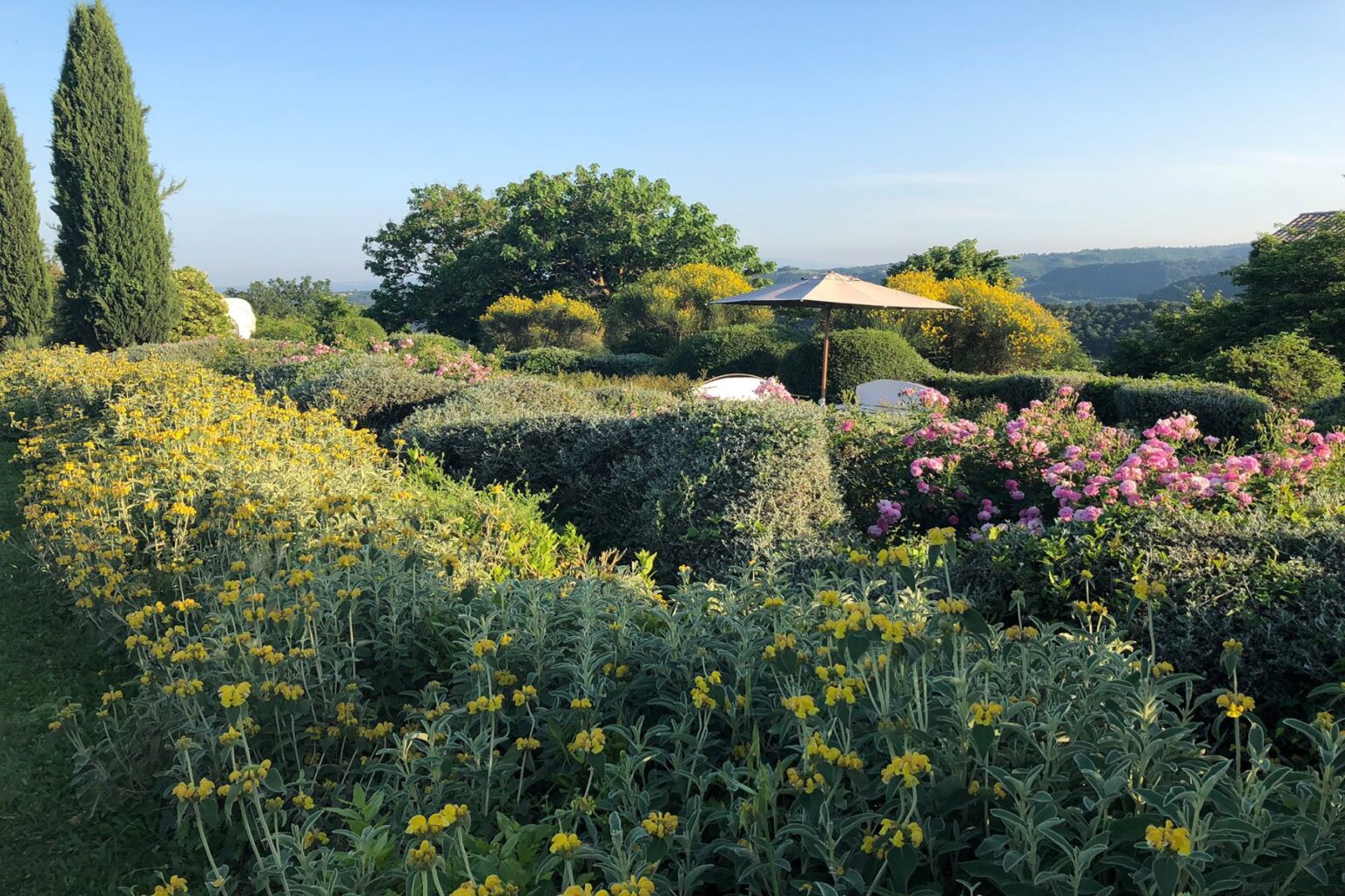 Lush garden landscape featuring vibrant yellow and pink flowers, neatly trimmed hedges, and a shaded seating area under an umbrella, set against a clear blue sky and rolling hills in the background. | Only on SopranoVillas