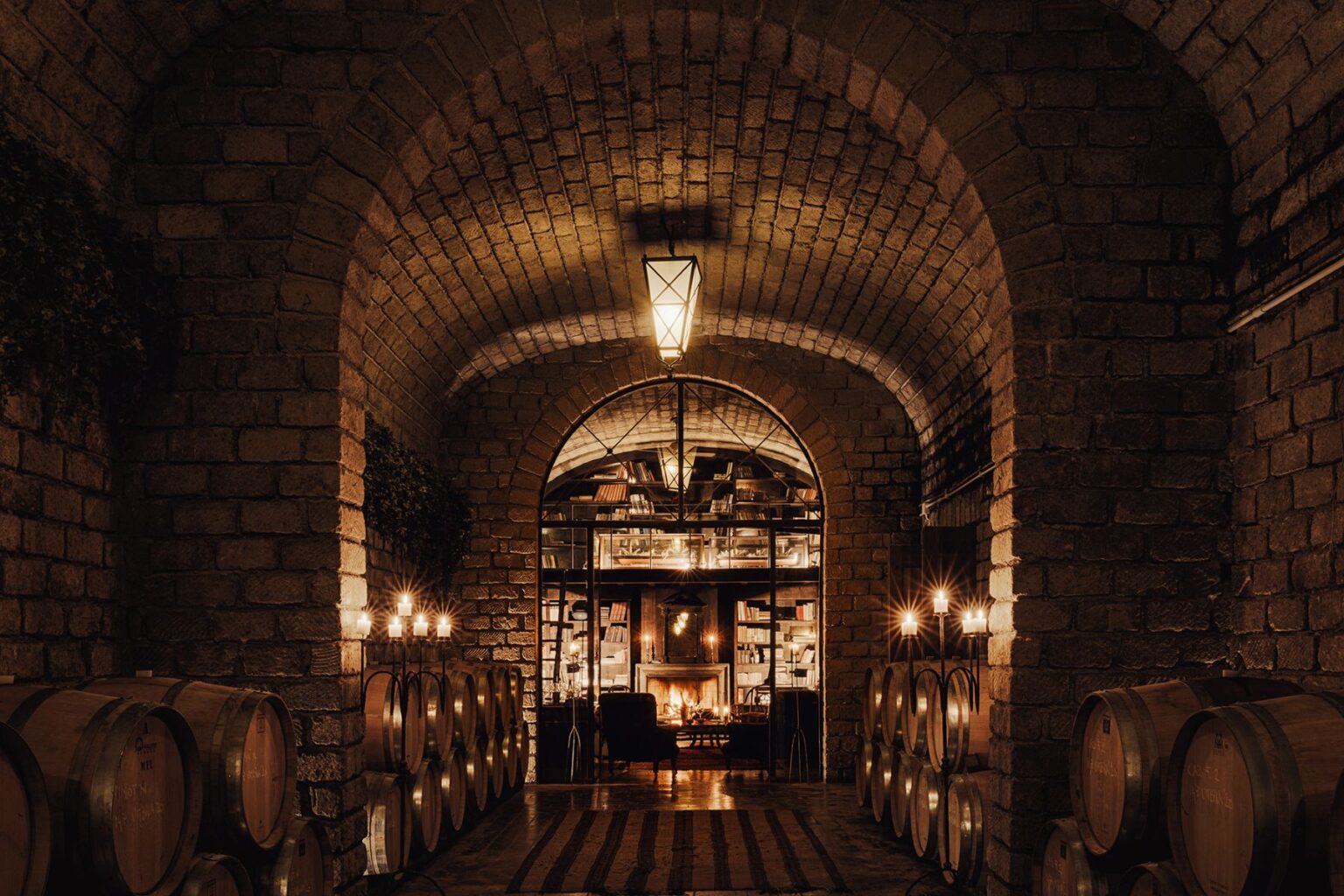 Rustic wine cellar interior featuring stone arches, wooden barrels, and warm candlelight, creating an inviting atmosphere with a cozy seating area and a fireplace in the background. | Only on SopranoVillas