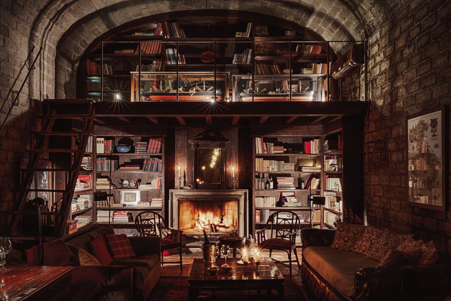 Cozy library interior featuring a stone wall, wooden bookshelves filled with books, and a warm fireplace. The space includes comfortable seating and a staircase leading to a second level, illuminated by candles and soft lighting, creating an inviting atmosphere for reading and relaxation. | Only on SopranoVillas