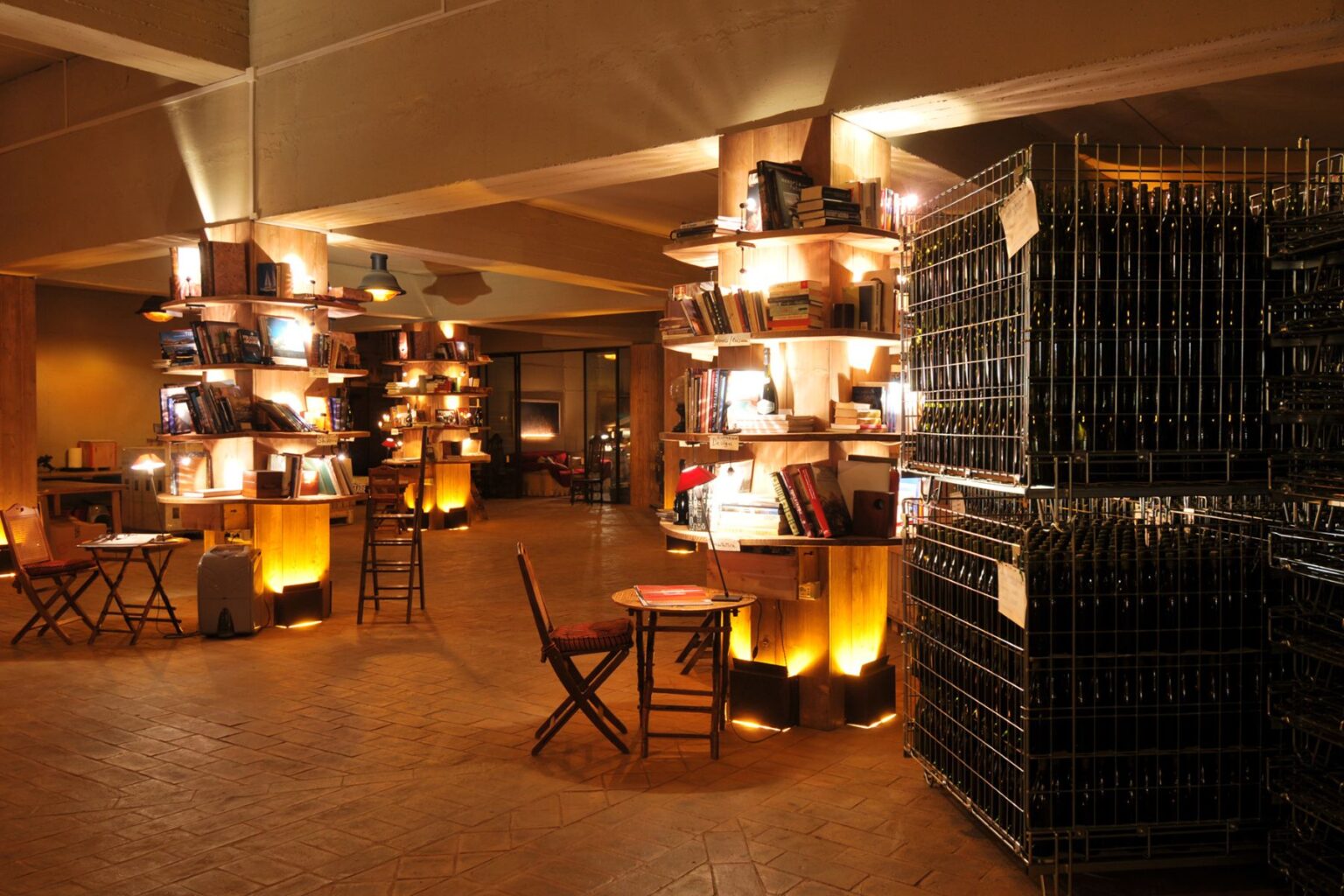 Cozy interior of a modern library featuring wooden shelves filled with books, warm lighting, and a small seating area with tables and chairs. A wine storage area is visible in the background, adding to the inviting atmosphere. | Only on SopranoVillas