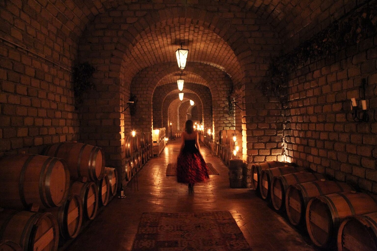 A woman in a flowing red dress walks through a dimly lit wine cellar with arched stone walls, lined with wooden barrels and flickering candles, creating an atmospheric and enchanting setting. | Only on SopranoVillas
