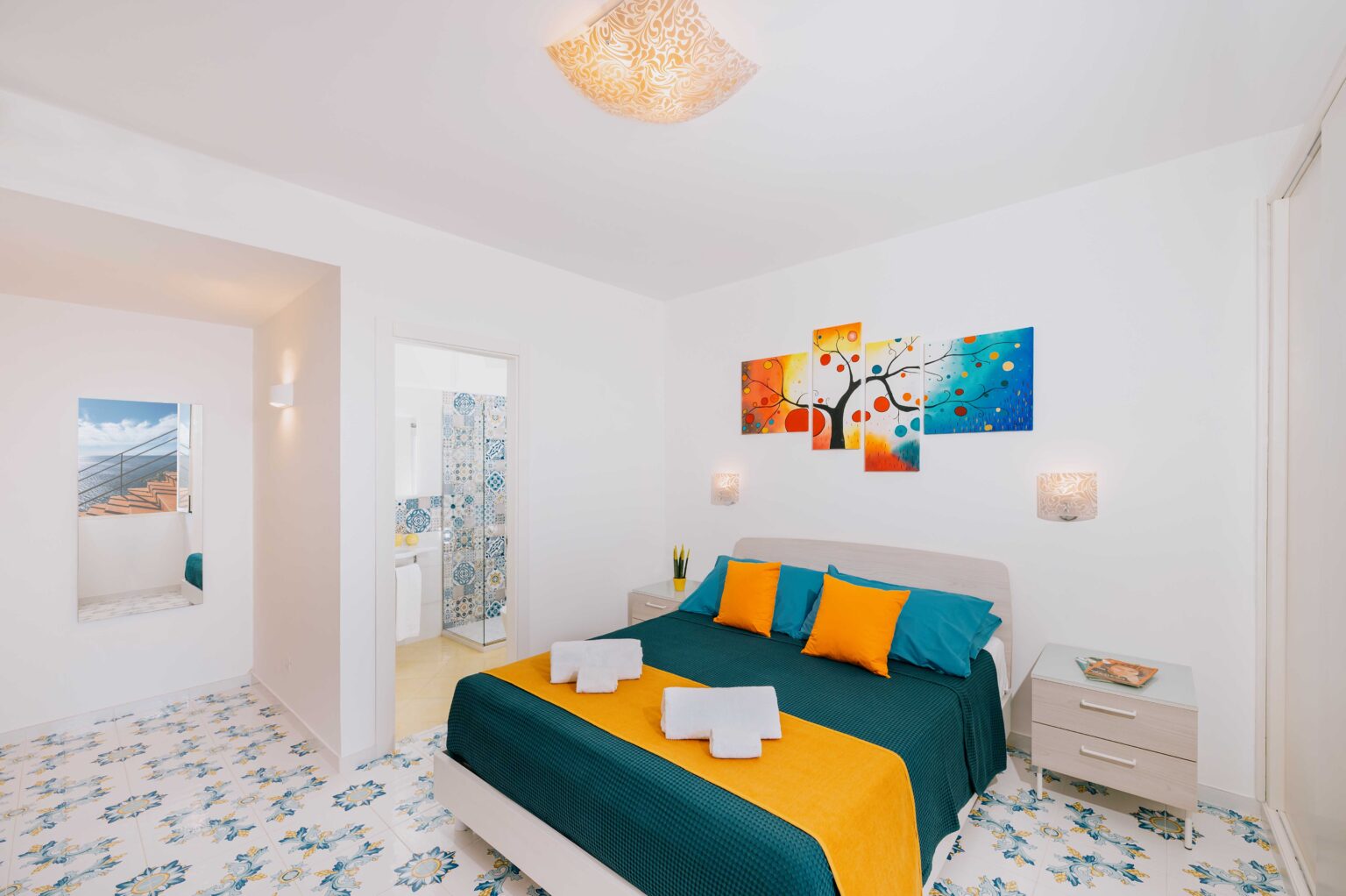 Bright and modern bedroom featuring a comfortable bed with vibrant blue and orange bedding, decorative wall art, and stylish lamps. The room has white walls and patterned flooring, with an adjacent bathroom visible in the background, creating a welcoming and relaxing atmosphere. | Only on SopranoVillas
