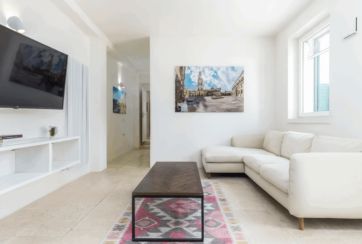 Modern living room featuring a white sectional sofa, a coffee table on a patterned rug, a large flat-screen TV, and a colorful wall art piece. Bright, airy space with natural light streaming through windows. | Only on SopranoVillas