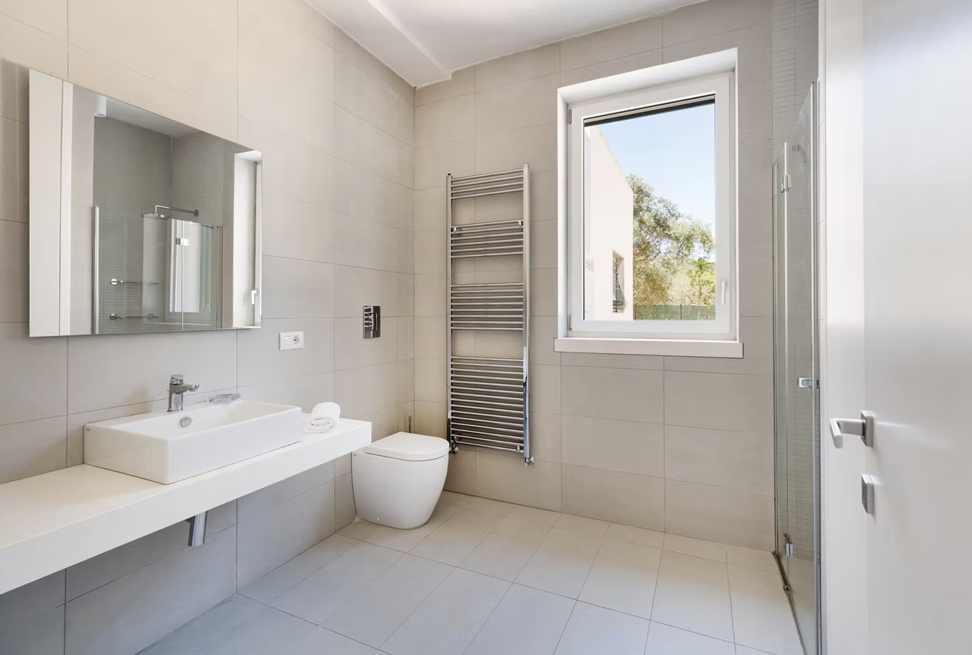 Modern bathroom featuring sleek design elements, a wall-mounted sink with a large mirror, a white toilet, and a towel radiator. Natural light streams in through a window, enhancing the spacious feel of the room. | Only on SopranoVillas