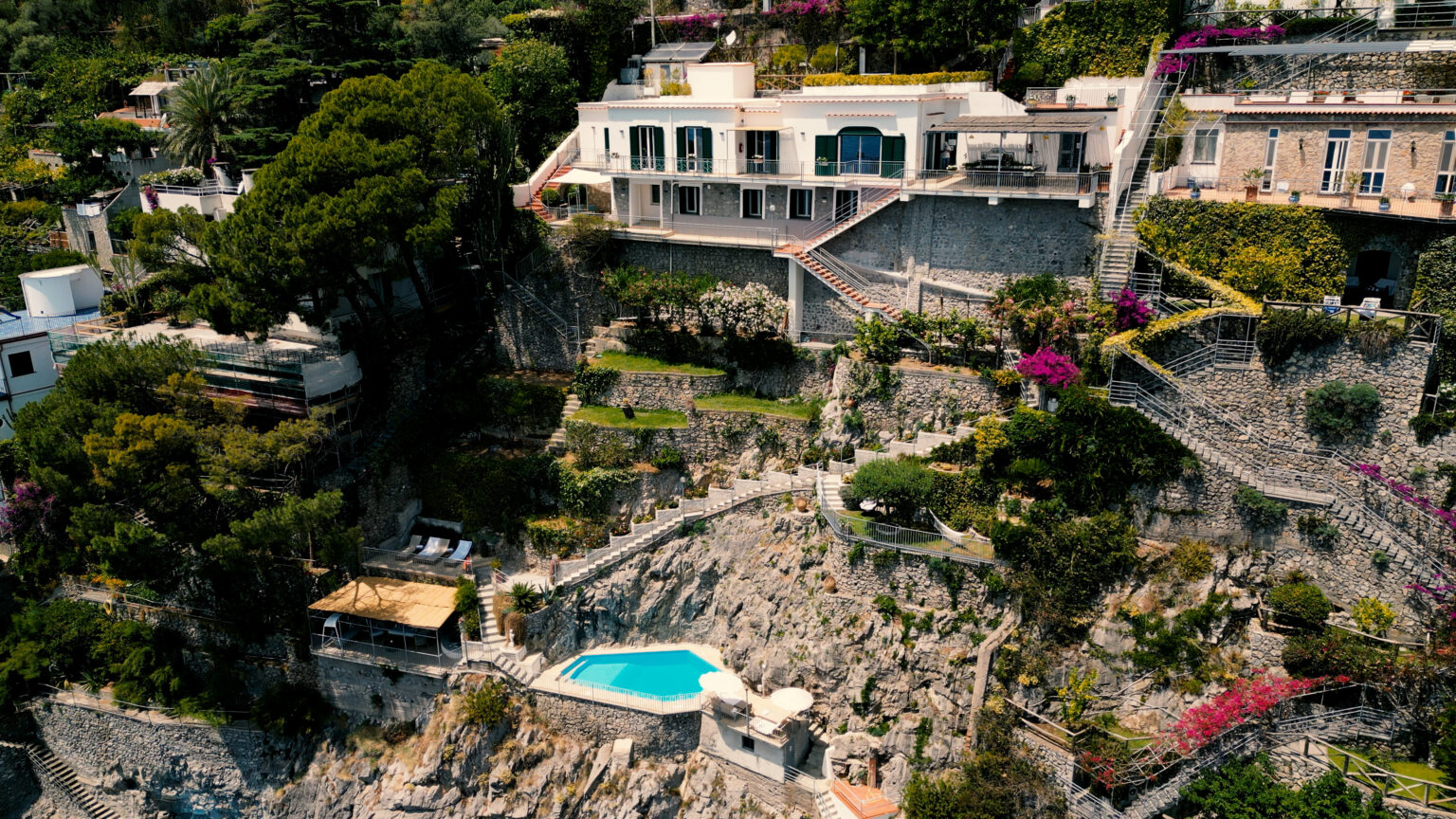 Aerial view of a luxurious hillside villa featuring a private pool, surrounded by lush greenery and vibrant flowers, overlooking a rocky landscape. | Only on SopranoVillas
