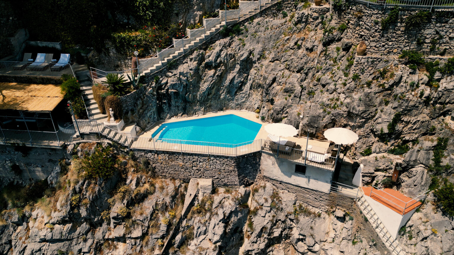Aerial view of a luxurious infinity pool situated on a rocky cliffside, surrounded by sun loungers and umbrellas, offering a serene retreat with stunning natural scenery. | Only on SopranoVillas