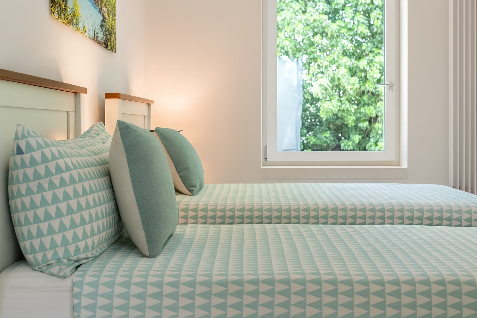 Luxurious bedroom interior in a villa for rent in Italy, featuring two single beds with stylish mint green patterned bedding and decorative cushions, complemented by natural light from a large window overlooking lush greenery. | Only on SopranoVillas