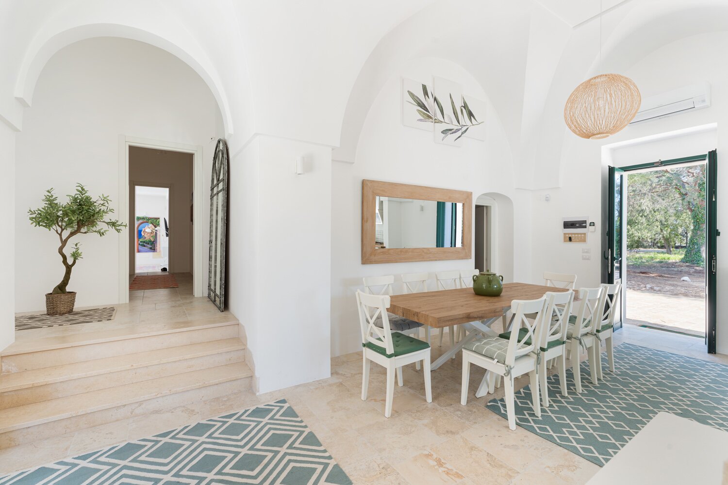Bright and airy dining area featuring a wooden table with white chairs, surrounded by large windows and an open doorway. The space is decorated with a potted plant, a large mirror, and a stylish light fixture, showcasing a modern yet inviting design. | Only on SopranoVillas