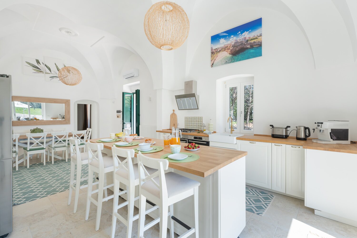 Bright and modern kitchen with white cabinetry, wooden countertops, and a dining area featuring a large table and chairs. The space is decorated with natural light, pendant lighting, and coastal artwork, creating a fresh and inviting atmosphere. | Only on SopranoVillas