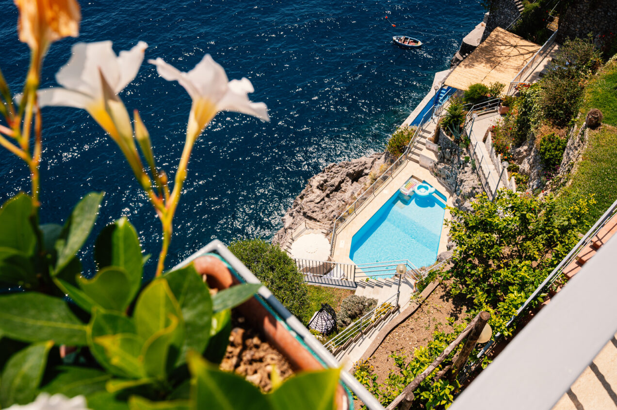 Aerial view of a luxurious coastal villa with a swimming pool overlooking the ocean, surrounded by lush greenery and blooming flowers. A small boat is visible on the water. | Only on SopranoVillas
