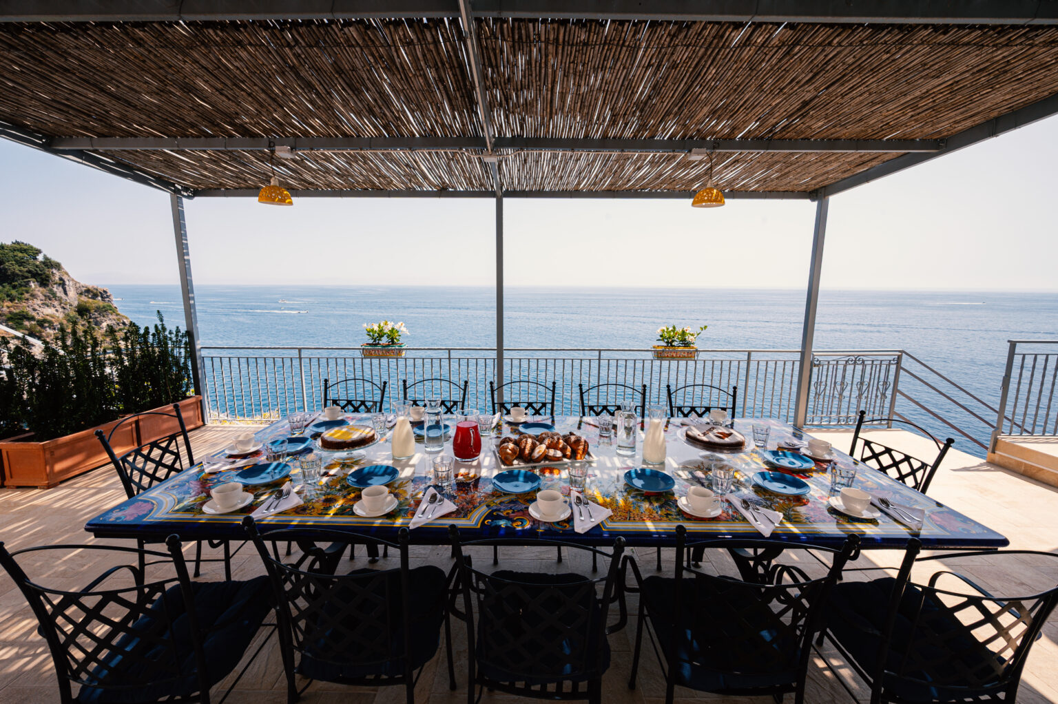 Outdoor dining table set for a meal, featuring blue plates, glassware, and a scenic ocean view under a thatched roof. Ideal for alfresco dining and gatherings. | Only on SopranoVillas