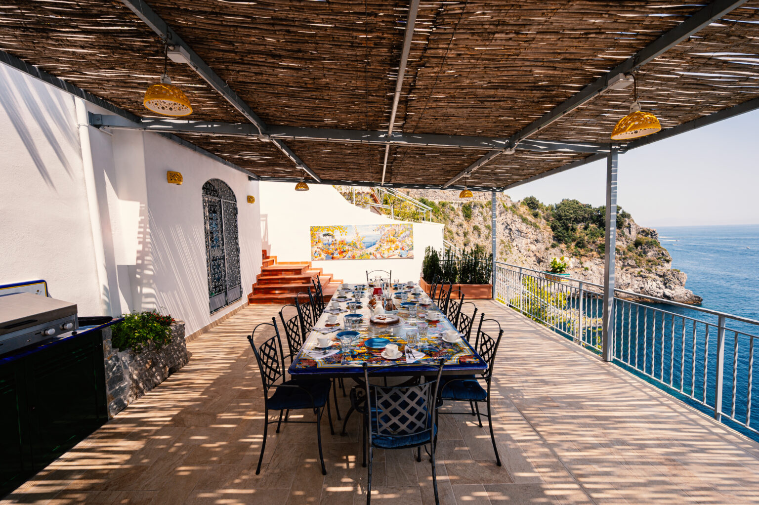 Outdoor dining area with a long table set for a meal, located on a terrace overlooking a scenic coastline. The space features a woven roof for shade, colorful table settings, and beautiful ocean views, creating an inviting atmosphere for gatherings. | Only on SopranoVillas