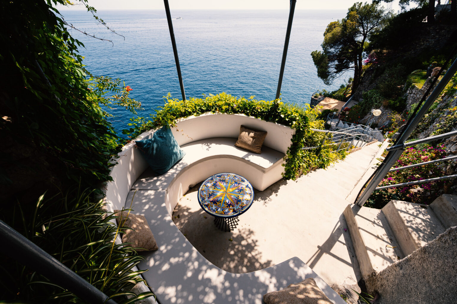 Cozy outdoor seating area overlooking the ocean, featuring a curved bench, colorful mosaic table, and lush greenery, perfect for relaxation and enjoying scenic views. | Only on SopranoVillas