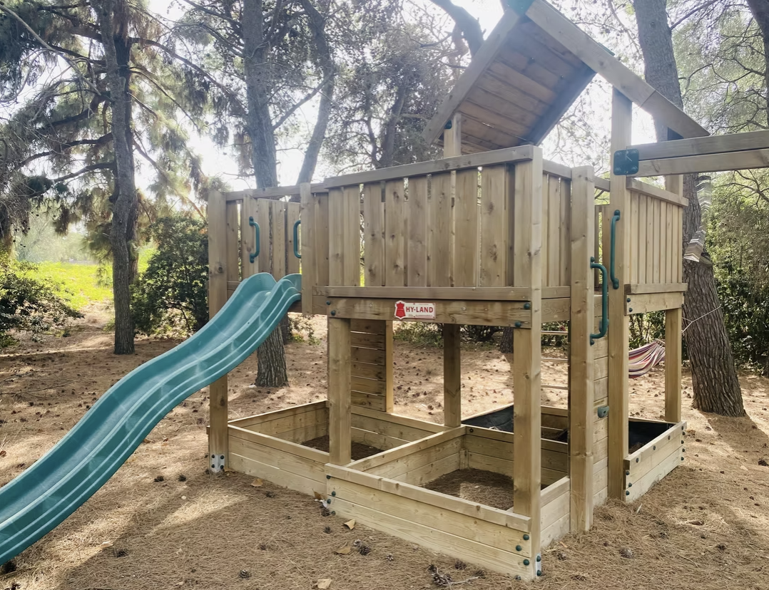 Wooden playset with a green slide and sandbox area, surrounded by trees in a backyard setting. Ideal for children's outdoor play and recreation. | Only on SopranoVillas