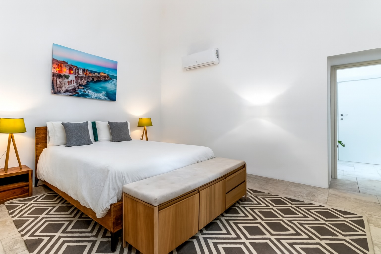 Modern bedroom featuring a large bed with white bedding, stylish lamps, a decorative rug, and an air conditioning unit, complemented by a vibrant wall art piece depicting a coastal scene. | Only on SopranoVillas