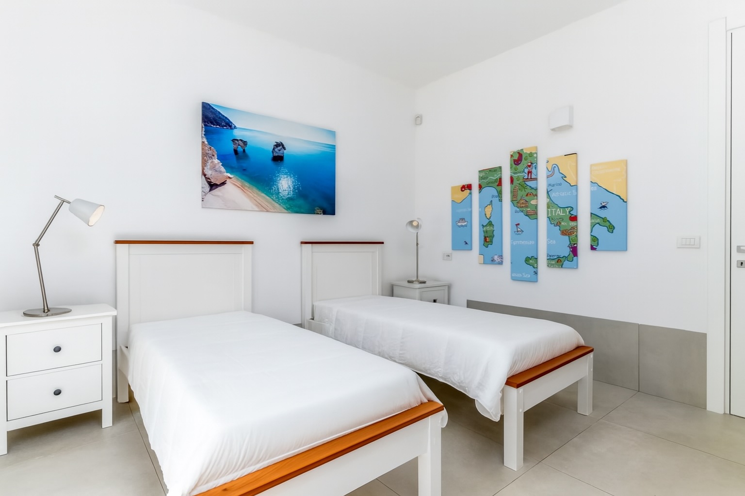 Bright and modern bedroom featuring two twin beds with white linens, a bedside table with a lamp, and a colorful wall art depicting maps of Italy. A large TV mounted on the wall adds contemporary flair to the space. | Only on SopranoVillas