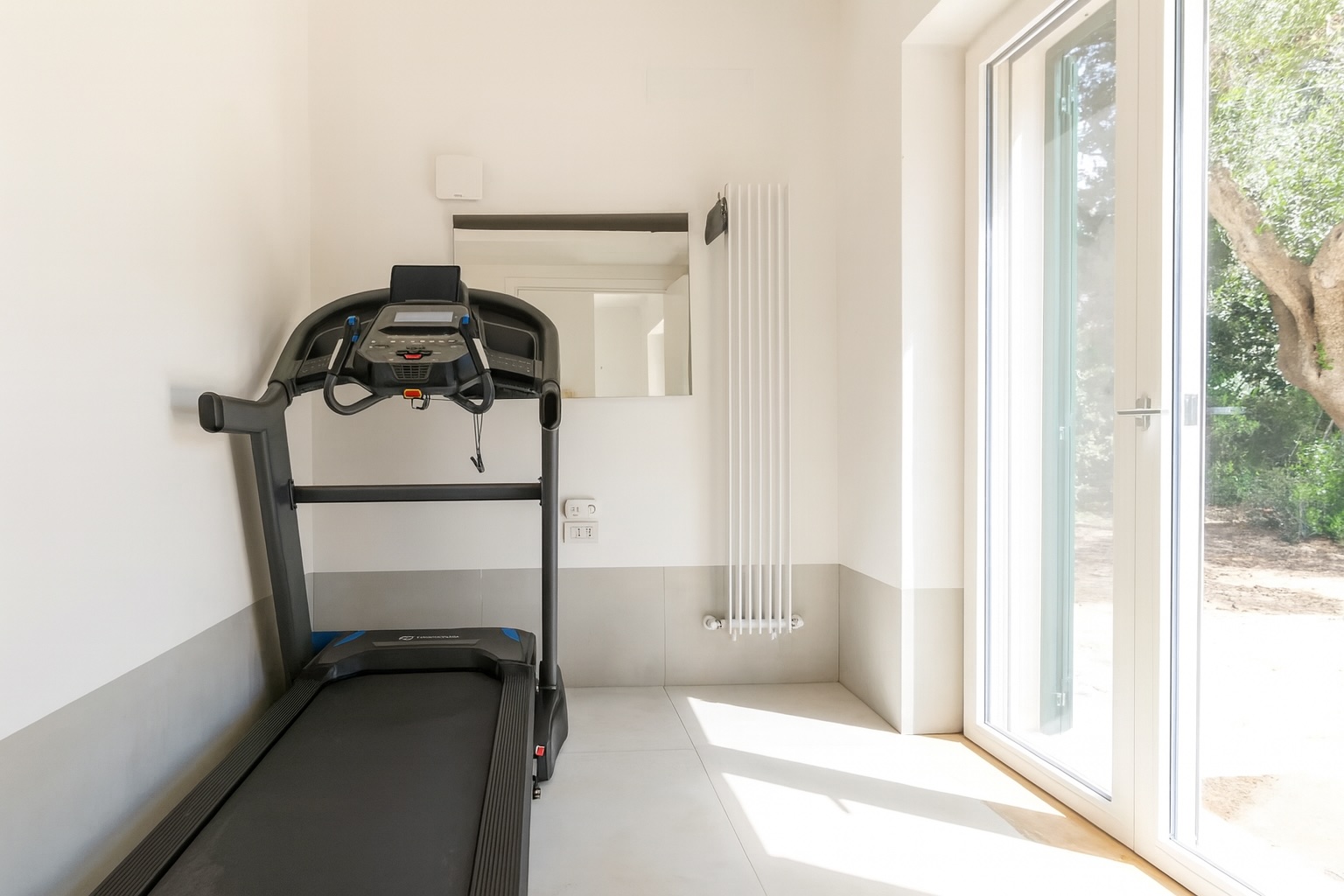 Luxury villa rental in Italy featuring a modern home gym with a treadmill, large windows allowing natural light, and a minimalist design for a tranquil workout space. | Only on SopranoVillas