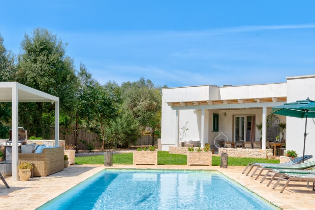Modern villa with a swimming pool surrounded by lush greenery, featuring outdoor seating and a shaded area with an umbrella, ideal for relaxation and outdoor living. | Only on SopranoVillas