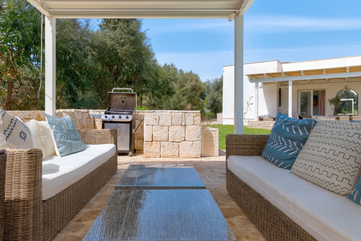 Luxurious outdoor seating area of a villa in Italy, featuring comfortable sofas with decorative pillows, a modern grill, and a scenic garden view, perfect for relaxing and entertaining guests. | Only on SopranoVillas