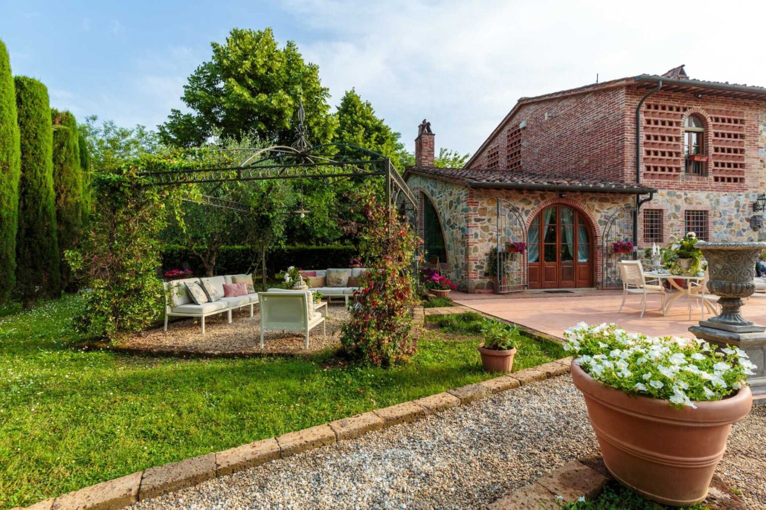 Charming outdoor patio area featuring a cozy seating arrangement under a decorative pergola, surrounded by lush greenery and blooming flowers, with a rustic stone house in the background. Perfect for relaxing and enjoying nature. | Only on SopranoVillas