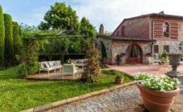 Charming outdoor patio area featuring a cozy seating arrangement under a decorative pergola, surrounded by lush greenery and blooming flowers, with a rustic stone house in the background. Perfect for relaxing and enjoying nature.