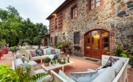 Cozy outdoor seating area with plush sofas and colorful cushions in front of a rustic stone house, surrounded by vibrant flowers and greenery, creating an inviting atmosphere for relaxation and social gatherings.