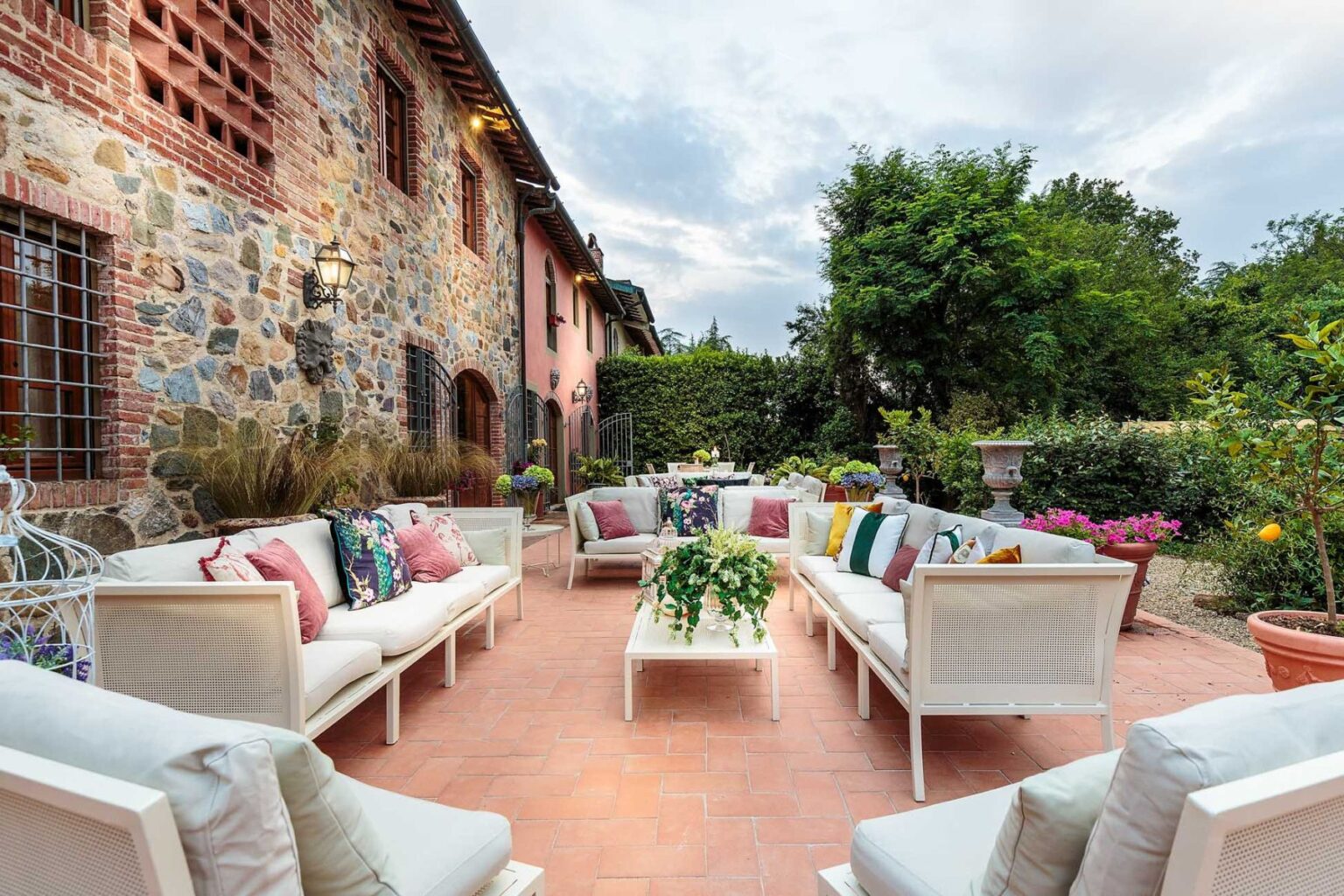 Cozy outdoor seating area featuring white sofas adorned with colorful cushions, surrounded by lush greenery and stone walls of a rustic building, ideal for relaxation and social gatherings. | Only on SopranoVillas