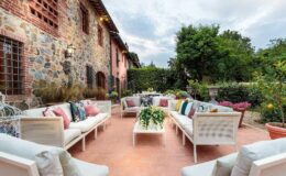 Cozy outdoor seating area featuring white sofas adorned with colorful cushions, surrounded by lush greenery and stone walls of a rustic building, ideal for relaxation and social gatherings.